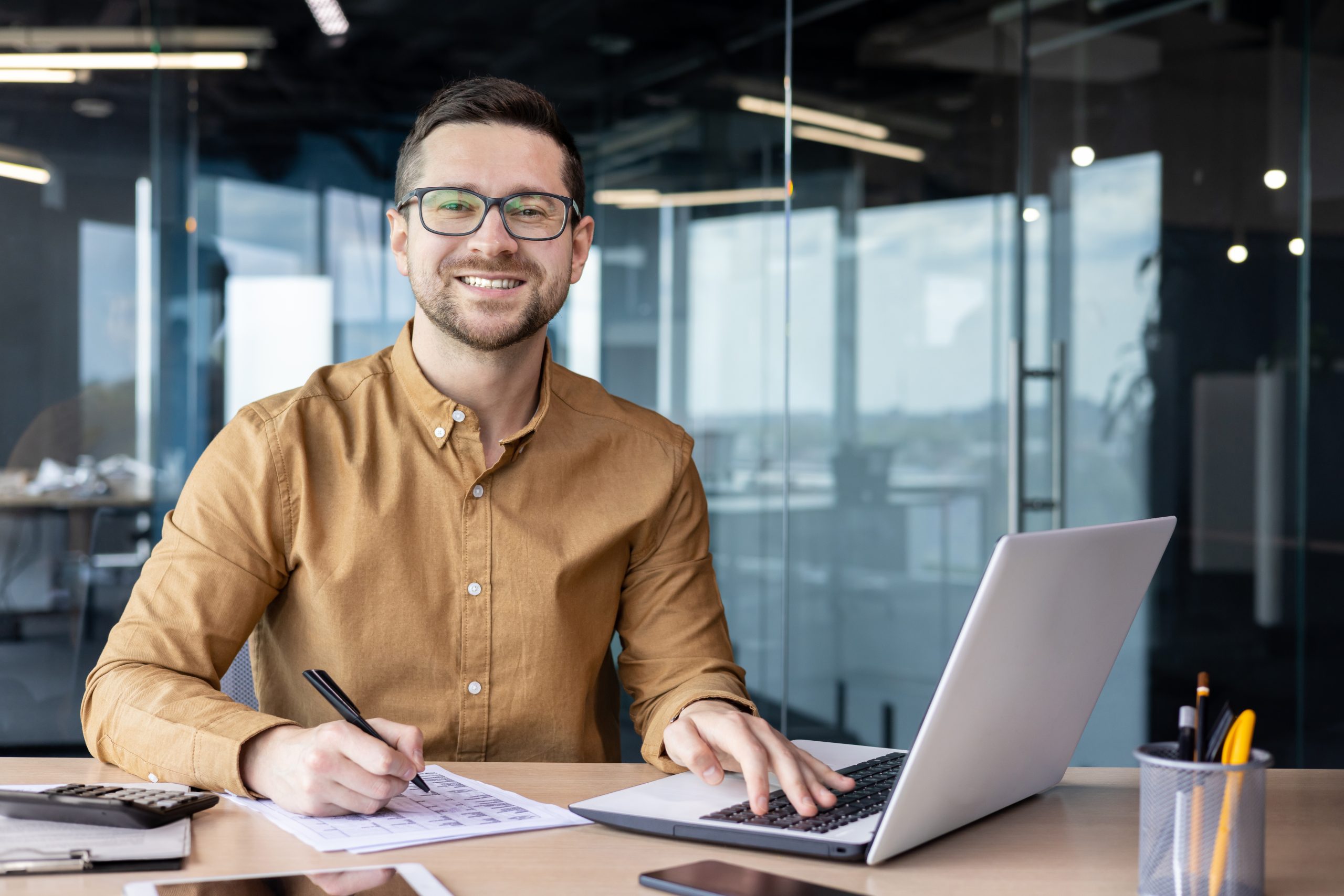 Portrait of young businessman in shirt, man smiling and looking at camera at workplace inside office, accountant with calculator behind paper work signing contracts and financial reports Portrait of young businessman in shirt, man smiling and looking at camera at workplace inside office, accountant with calculator behind paper work signing contracts and financial reports
